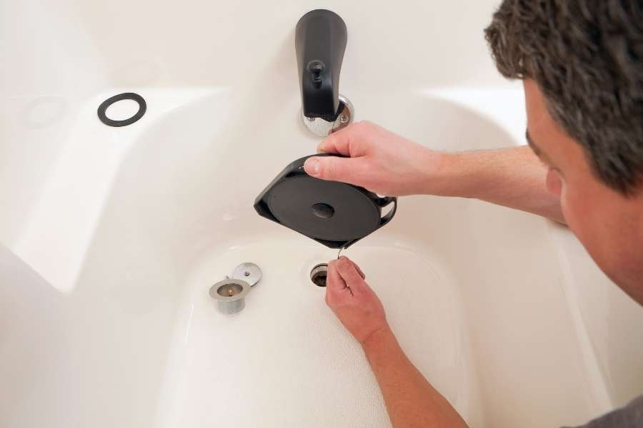 A man uses a plastic drain snake tool to clear hair and debris from a bathtub drain.